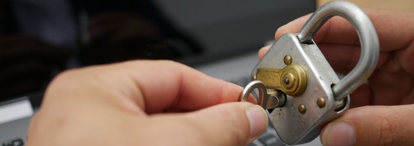 Eine Hand die ein Vorhängeschloss Hält. Eine andere Hand steckt einen Schlüssel in das Schloss. Im Hintergund ist ein Laptop zu sehen.